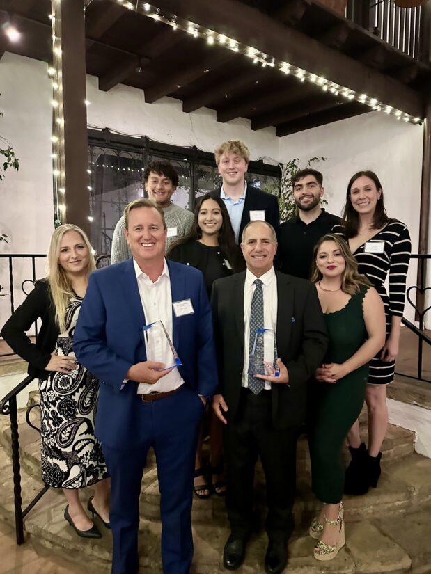 A group photo of Maho Prentice, LLP attorneys and staff at an award event, featuring two attorneys in front holding mentorship award plaques, surrounded by smiling team members in a formal setting with string lights.